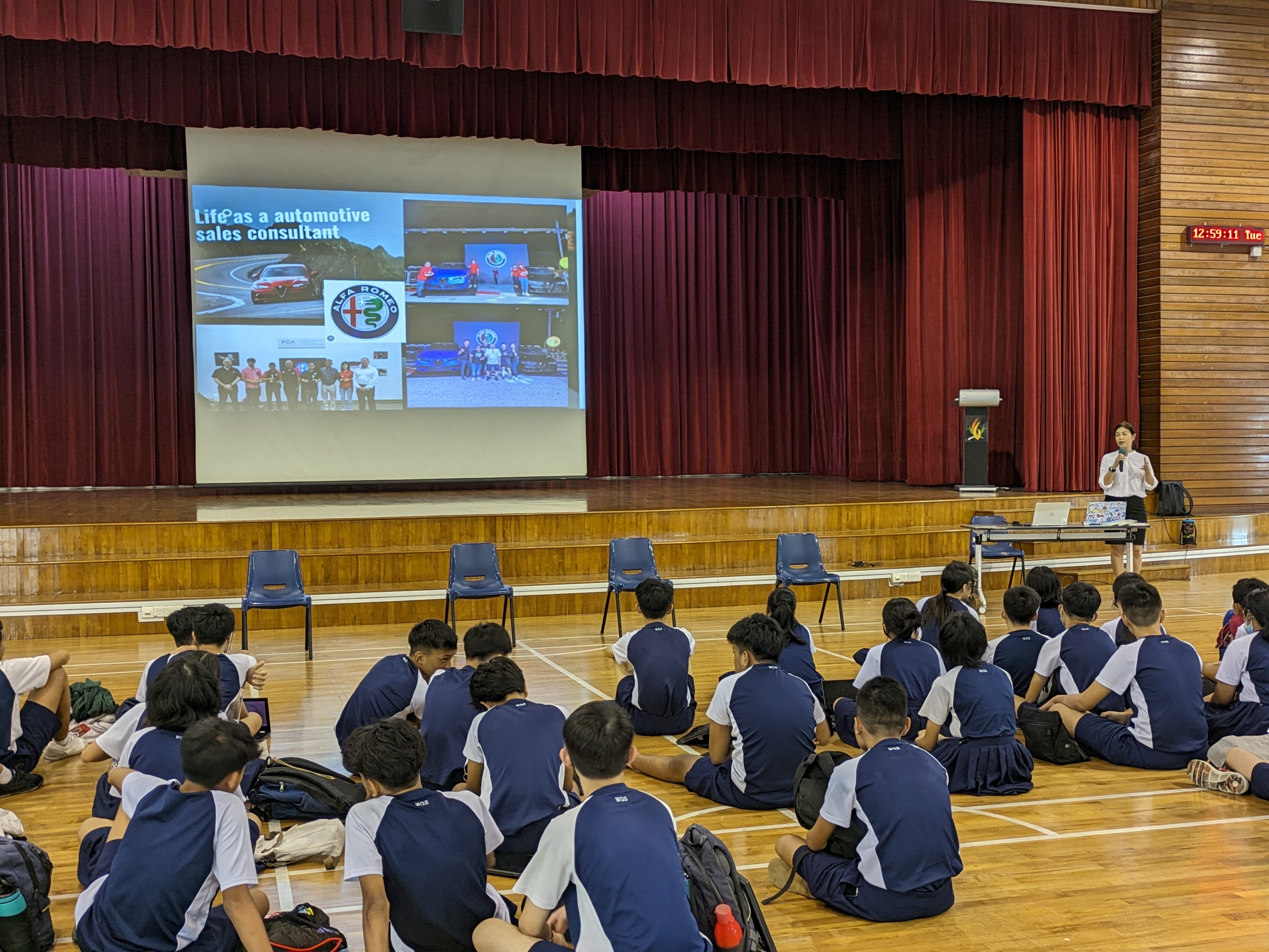 Students in uniforms sit on a hall floor watching a presenter speak beside an Alfa Romeo automotive sales consultant slideshow.